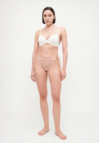Bralette blanche avec un design en V et des fines bretelles, associée à un string en dentelle rose clair avec des motifs en forme de cœur et des côtés à nouer.
