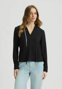 Zwarte top met een V-hals, voorzien van lange mouwen en een decoratieve strook met zilveren accenten aan één zijde. Glad stof, minimalistisch ontwerp.