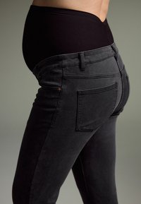Trudnički traperice u crnom denimu imaju visoki struk s mekanim materijalom za udobnost, uski kroj i stražnje džepove. Teksturirana površina poboljšava elastičnost.