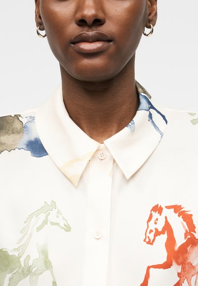 Lähikuva henkilöstä, joka pitää valkoista kauluksellista paitaa, jossa on vihreän ja punaisen vesivärillä maalattuja hevosen kuvia kankaalla.