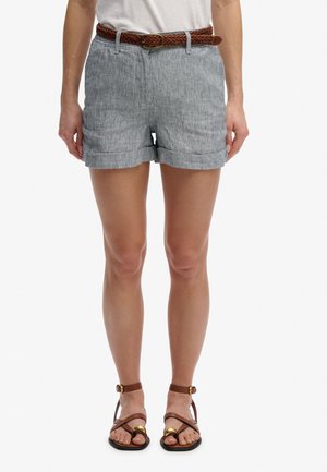 Kvinde iført gråstribede shorts med et brunt flettet bælte og brune sandaler med ankelrem, stående foran en ensfarvet hvid baggrund.