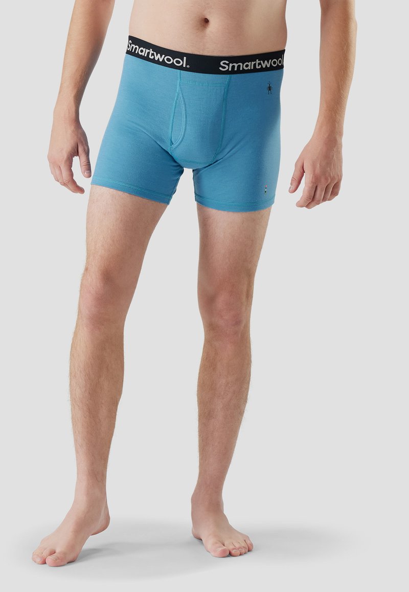 Homme debout pieds nus portant un boxer Smartwool bleu avec une ceinture élastique noire à logo, sur un fond uni.