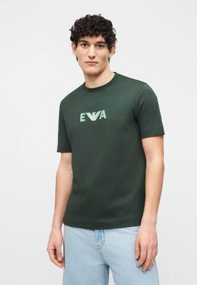 Νέος άνδρας με σγουρά μαλλιά που φοράει κοντομάνικη μπλούζα σκούρου πράσινου χρώματος με το λογότυπο "EWA" και ανοιχτόχρωμο τζιν μπροστά σε απλό φόντο.