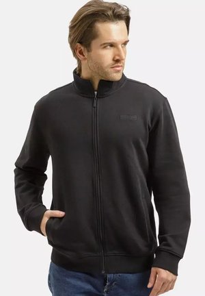 Mężczyzna w czarnej kurtce na zamek z wysokim kołnierzem i niebieskich dżinsach, patrzący na bok, z jedną ręką w kieszeni.