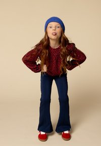 Rode bloemen lange mouwen top, blauwe gebreide beanie, marineblauwe flared broek en rode schoenen. De figuur maakt een speelse pose met een tonggebaar.