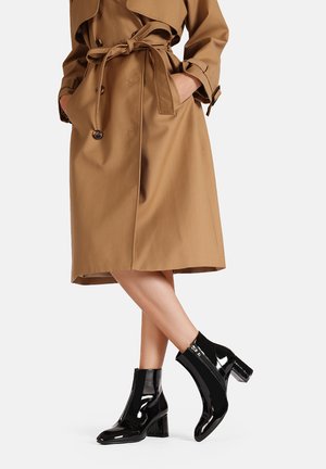 Stivali alla caviglia in pelle verniciata nera con tacco a blocco, abbinati a un trench coat color cammello con cintura annodata e grandi bottoni.