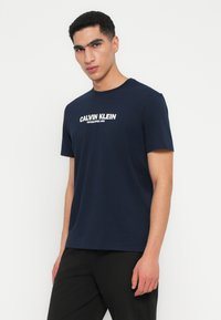 Tumši zils kokvilnas T-krekls ar apaļu kakla izgriezumu un baltu teksta logotipu "CALVIN KLEIN ESTABLISHED 1968" uz krūšu daļas. Īsas piedurknes.