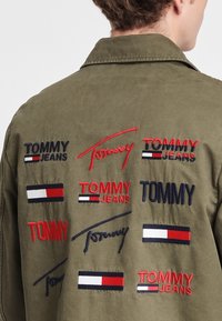 Olive green jacket with multiple embroidered logos in red, navy, and white, featuring a smooth texture and a button-up design.