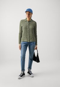 Olivovozelená textúrovaná košeľa na gombíky, modrá čiapka, modré džínsy, čierne tenisky a čierna kabelka. Neformálny outfit s rôznymi materiálmi.
