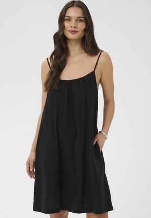 Mujer con cabello largo y castaño, vestida con un vestido negro sin mangas y suelto con tirantes delgados, una mano en el bolsillo, sobre un fondo liso.