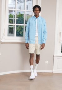 Camicetta azzurra chiara con bottoni, t-shirt bianca sotto, pantaloni corti beige, calze alte bianche con dettagli e sneakers bianche. Ambientazione interna.