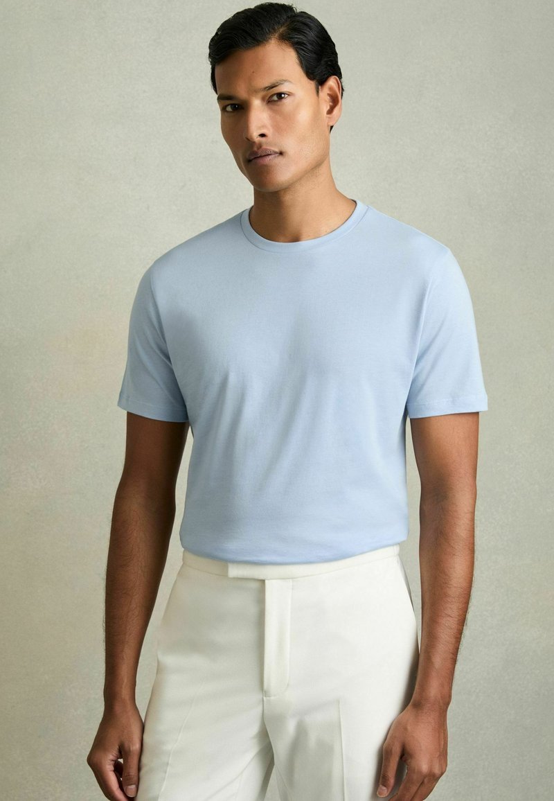 Tricou scurt cu mânecă scurtă de culoare albastru deschis, dintr-un material moale, cu guler rotund, asortat cu pantaloni albi, croiți, cu o textură netedă și un design simplu.