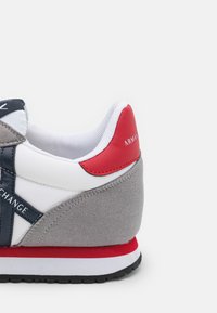 Sapatilha cinza com detalhes em branco e azul marinho, com um tabuleiro em vermelho e camurça texturizada. Logotipo em relevo e sola de borracha contrastante.