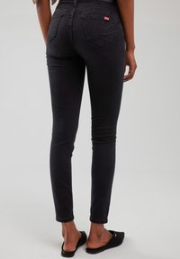 Žena u crnim uskim trapericama i crnim obućama na navlačenje, stoji okrenuta leđima prema kameri na jednobojnoj svijetloj pozadini.