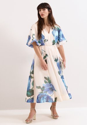 Femme portant une robe midi crème avec des imprimés floraux bleus, des manches papillon, une ceinture à la taille, tenant une pochette, et des sandales dorées à talons hauts.