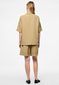Chemise beige à manches courtes et short assorti en tissu lisse. Le short présente des ourlets retroussés et une coupe décontractée, associé à des chaussures noires.