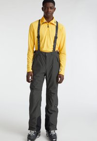 Pantalones de esquí en gris oscuro con tirantes negros ajustables, textura suave y bolsillos laterales ocultos. Se llevan con una blusa de manga larga amarilla.