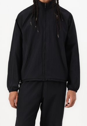 Personne portant une veste noire zippée et un pantalon noir assorti, debout devant un fond blanc uni.
