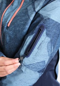 Giacca blu resistente all'acqua con motivi testurizzati, cerniere arancioni a contrasto e una tasca con zip sul braccio che presenta un logo.
