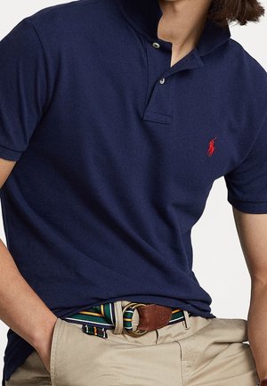 Polo bleu marine avec une patte de boutonnage à trois boutons, manches courtes, et logo rouge. Associé à une ceinture rayée multicolore et un pantalon kaki.