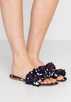 Bare feet wearing flat open-toe slippers with blue, white, and brown ruffled fabric straps on a smooth gray surface.