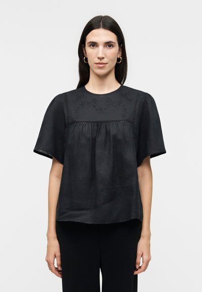 Femme aux longs cheveux foncés portant des boucles d'oreilles créoles en or et un chemisier noir ample à manches courtes larges avec une broderie florale sur le yoke.