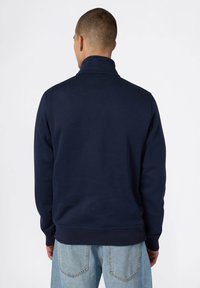 Pullover blu navy con colletto alto e polsini a costine. Tessuto morbido, vestibilità rilassata, abbinato a jeans di denim blu chiaro. Vista posteriore.
