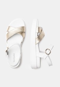 Sandales blanches avec une semelle épaisse, dotées d'un design croisé en brides métalliques dorées et d'une sangle de cheville ajustable avec une boucle dorée.