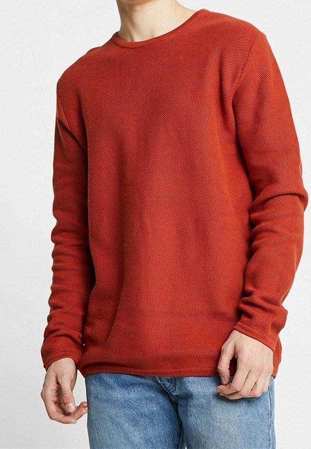 Red textured knit sweater with a crew neckline and long sleeves, styled with blue jeans, showcasing a relaxed fit and ribbed cuffs.