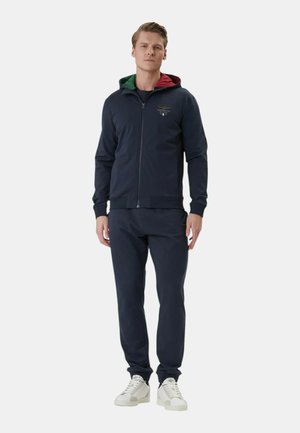 Giovane uomo in piedi che indossa una felpa con cappuccio blu navy con zip e fodera rossa e verde, pantaloni coordinati e sneakers bianche su sfondo semplice.