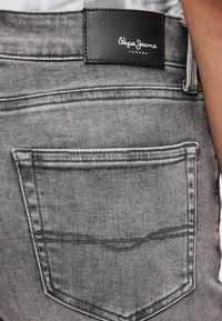 Szare dżinsy z denimu z tylną kieszenią i czarną skórzaną metką marki z białym logo. Widoczne subtelne przeszycia wzdłuż szwów.