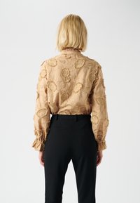 Beige embroidered blouse with floral patterns, long sleeves, and ruffled cuffs, paired with black tailored pants.