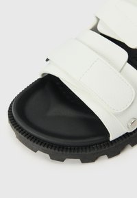 Sandal in pelle bianca con due cinturini regolabili, superficie testurizzata e suola in gomma nera con un motivo segmentato e bordi rialzati.