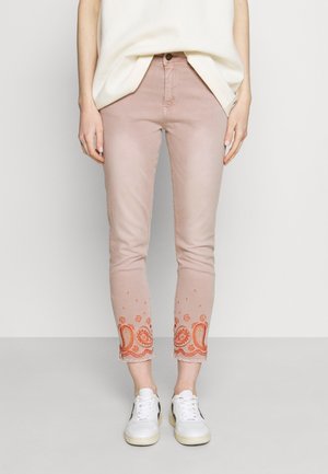Femme portant un pantalon slim rose clair avec une broderie paisley orange aux ourlets raccourcis et des baskets blanches, debout devant un fond uni.