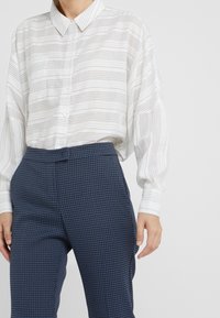 Wit gestreepte blouse met lange mouwen en knoopsluiting, gecombineerd met blauwe broeken met een klein geometrisch patroon.