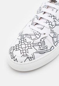 Witte sneaker met zwarte geometrische en bloemmotieven, witte veters en een getextureerde, witte rubberen zool, gefotografeerd op een effen witte achtergrond.