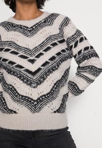 Personne portant un pull en tricot doux avec des motifs chevrons et rayures noirs et gris, assorti à un pantalon foncé, sur un fond uni.