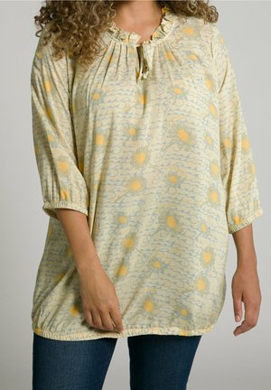 Krøllet-haired person iført lysegul bluse med blå kursiv tekst og gule solsikker, kombineret med mørkeblå jeans, står mod en ensfarvet baggrund.