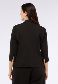 Czarna, dopasowana marynarka z rękawami 3/4 i bocznymi rozcięciami. Gładki materiał, stójka. Stylizowana z pasującymi czarnymi spodniami.