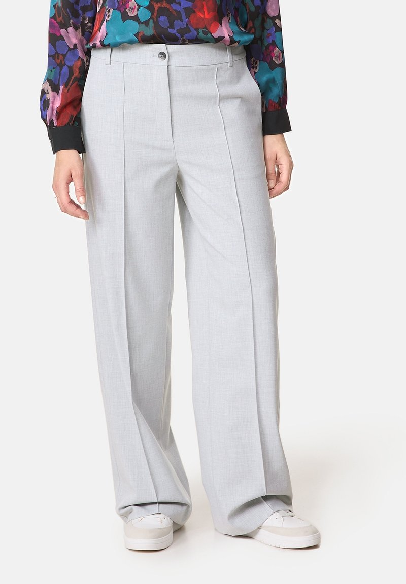 Personne portant un pantalon large gris clair avec des plis sur le devant, associé à des baskets blanches et un chemisier fleuri coloré à manches longues.