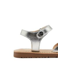 Sandali metallici argento con design a T-strap, parte superiore testurizzata e suola piatta marrone. Presentano un accento con bottone marchiato.