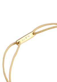 Bracelet plaqué or avec un charm rectangulaire lisse marqué "925 Ag," sur un cordon tressé beige. Présente des accents métalliques et un fermoir.