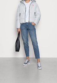 Světlá šedá péřová bunda se zipem, modré džíny a metalické stříbrné tenisky. Černá taška přes rameno. Jednoduché pozadí.