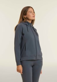 Felpa grigia scura con zip, colletto alto, cordini, tasche laterali e polsini a coste, realizzata in tessuto morbido e elasticizzato.