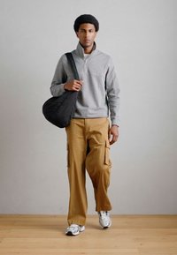 Γκρι φούτερ με φερμουάρ, μπεζ cargo παντελόνι με πλαϊνές τσέπες και γκρι αθλητικά παπούτσια. Το μοντέλο κρατά μια μαύρη στρογγυλή τσάντα ώμου.