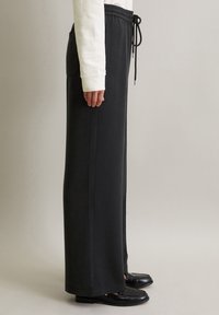 Personne portant un pantalon large noir avec des poches côtés et une taille à cordon, associé à des mocassins noirs et un haut à manches longues blanc.