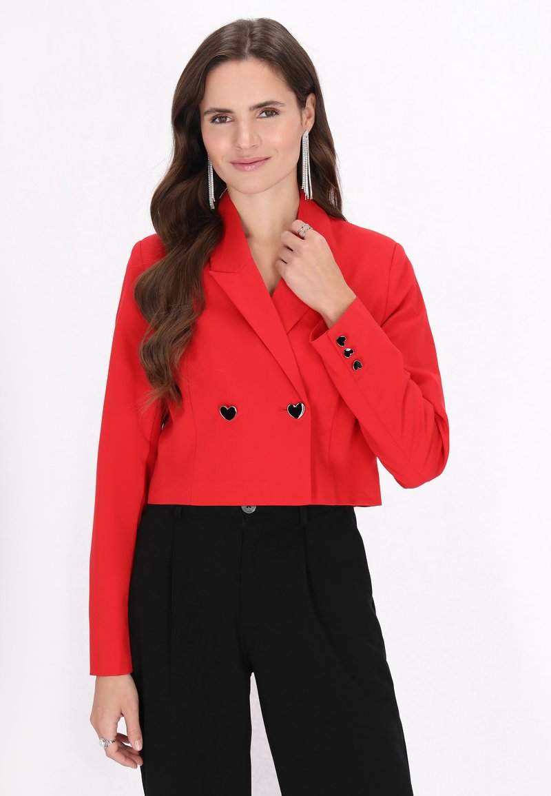 Blazer rosso cropped con design doppiopetto, bottoni a forma di cuore, maniche lunghe e tessuto morbido, abbinato a pantaloni neri.