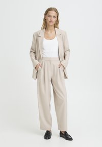 Woman standing with hands in pockets wearing beige suit, white top, and black loafers against a plain white background.