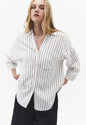 Mujer con cabello rubio largo, vistiendo una camisa blanca abotonada con finas rayas verticales y pantalones negros, posando con las manos detrás de la espalda.