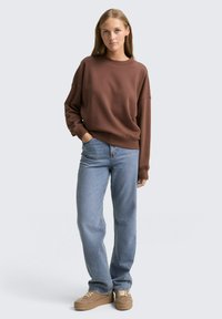 Brązowy, oversize'owy sweatshirt z okrągłym dekoltem, w połączeniu z jasnoniebieskimi prostymi dżinsami oraz beżowymi sneakersami na grubej podeszwie.